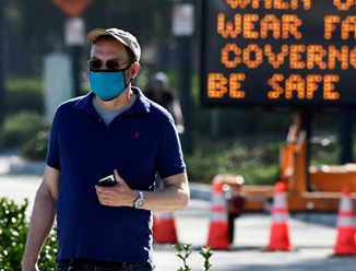 A pedestrian wearing a face mask walking in front of a sign that asks people to be safe